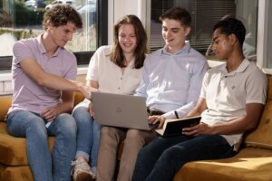 Een groep jonge marketing stagiaires bespreekt samen een project op een laptop, terwijl ze comfortabel zitten op een gele bank bij het raam. Ze zijn geconcentreerd en betrokken, wat duidt op een actieve samenwerking en het delen van ideeën tijdens hun marketing stage.